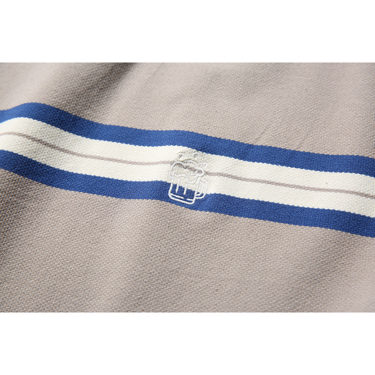 Close-up of a small white embroidered emblem on the taupe shirt's blue and ivory stripes.