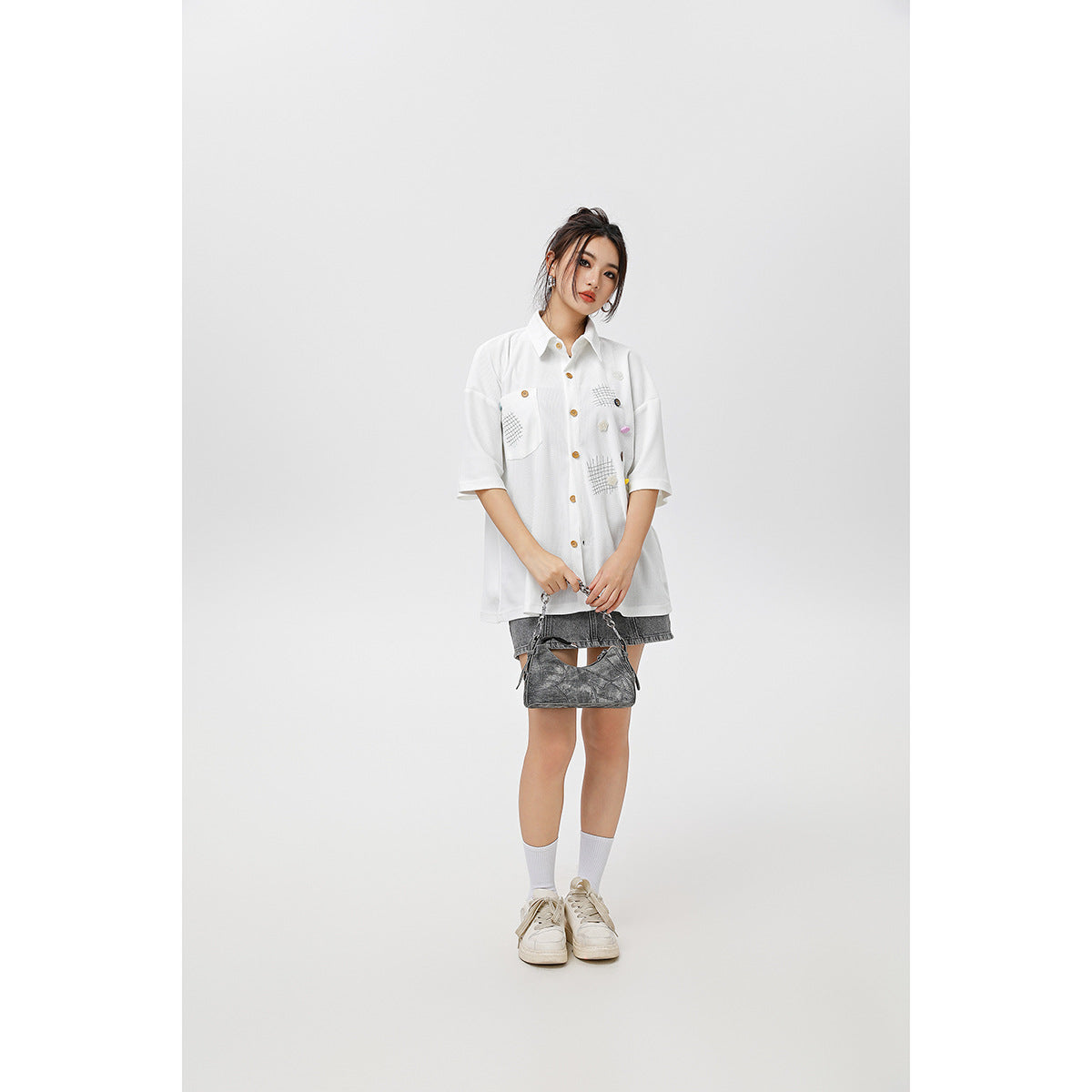 Model wearing an oversized white short-sleeve button-up shirt with grid patches and colorful accents, paired with a gray