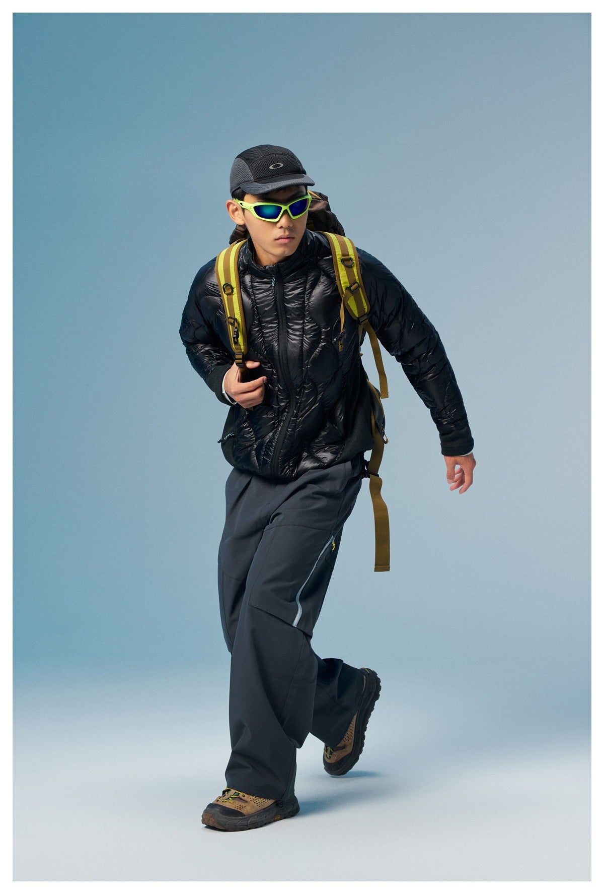 A man strides in a black quilted zip jacket, dark trousers, and a hiking backpack.