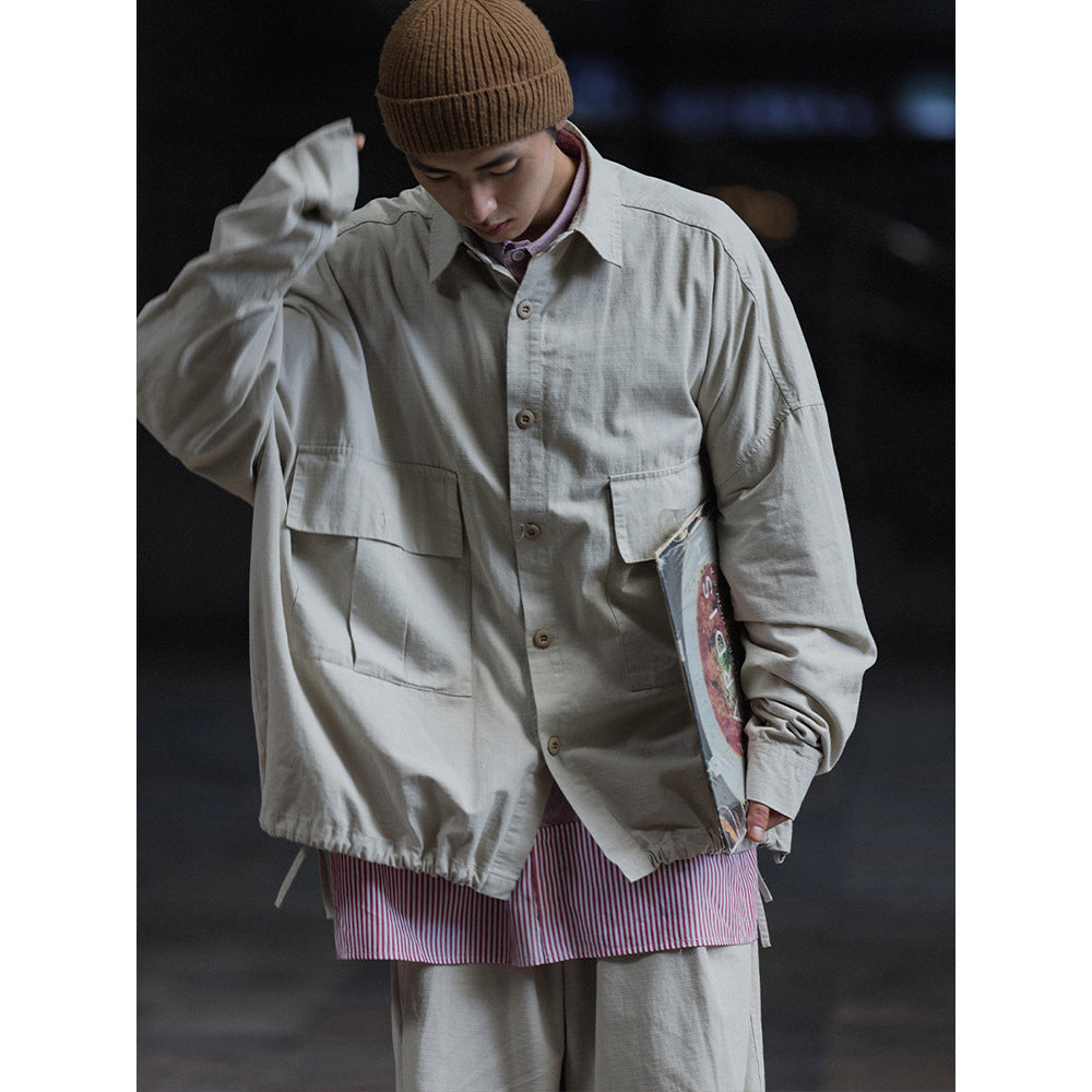 Model wearing a light gray oversized button-up jacket with large flap pockets, paired with gray trousers and a brown beanie.