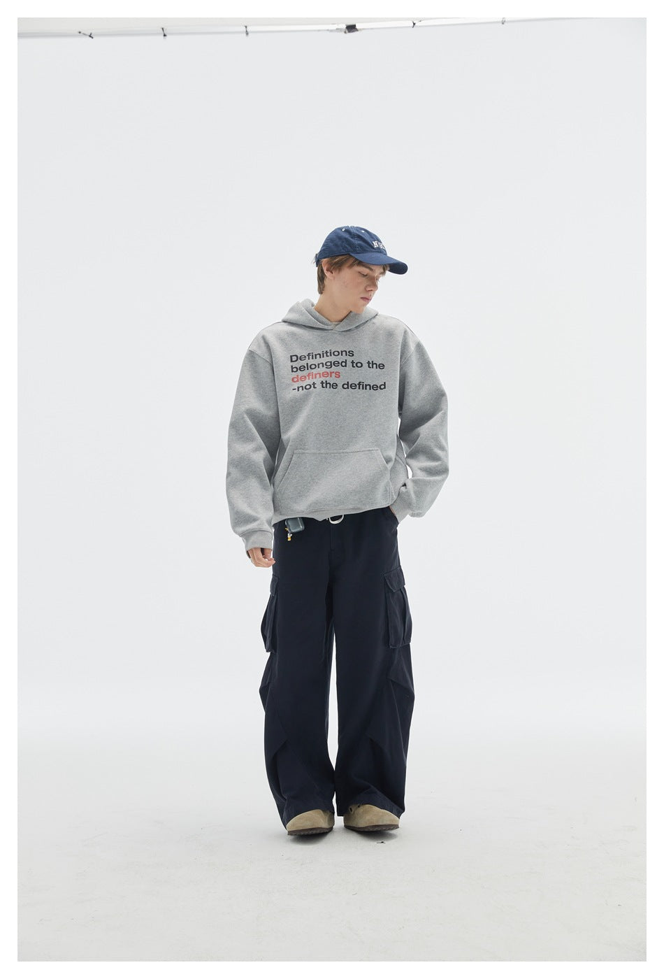 Model wearing a gray slogan-print pullover hoodie with dark cargo pants, shown full length.