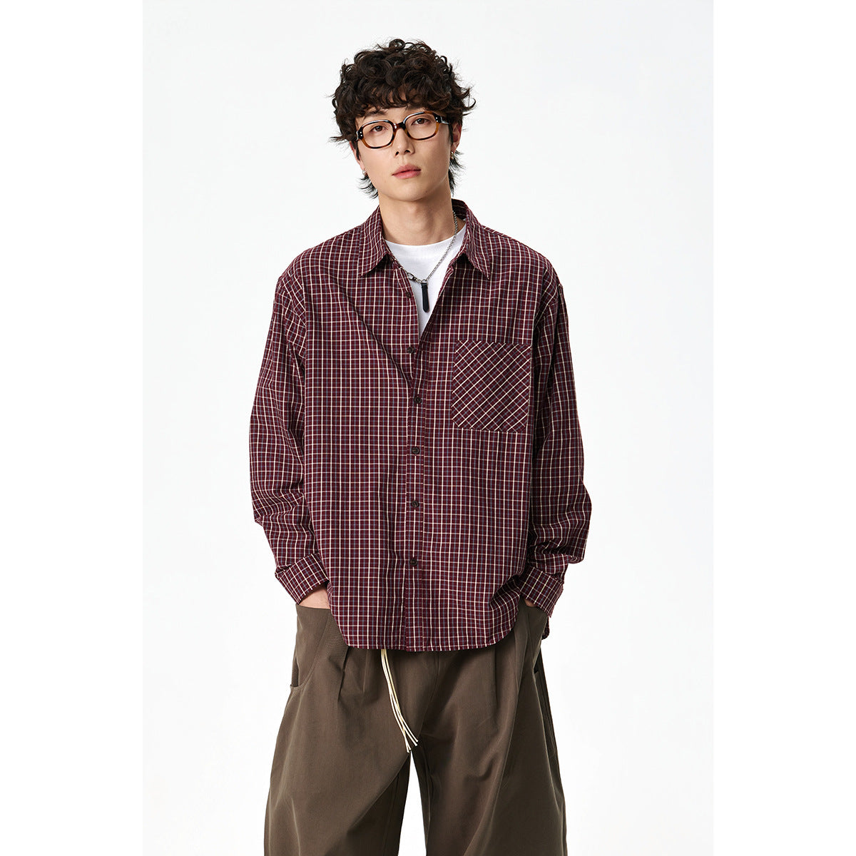 Front view of a model wearing a burgundy plaid button-up shirt with a contrasting chest pocket and loose brown trousers.