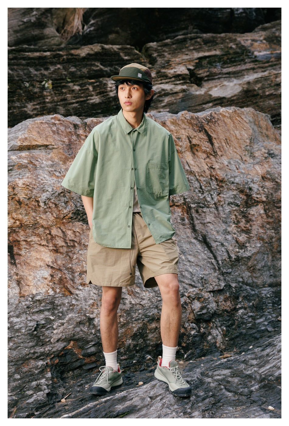 Model wearing a pale sage oversized short-sleeve button-up shirt with a chest pocket and tan shorts outdoors.
