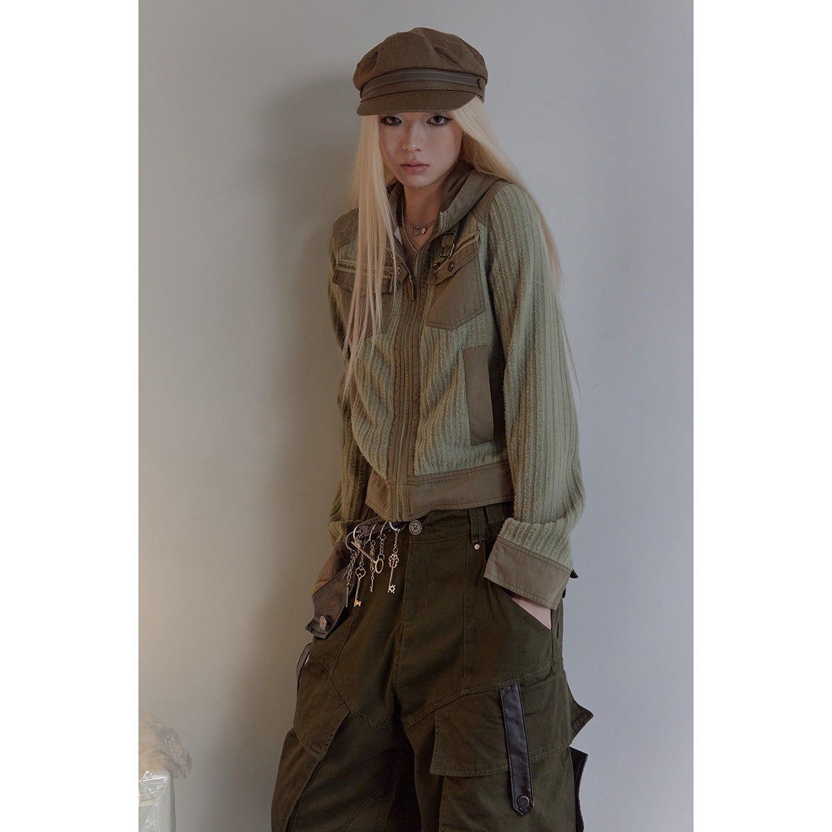 Model wearing a sage ribbed zip jacket with tan panels, dark olive cargo pants, and a brown cap.