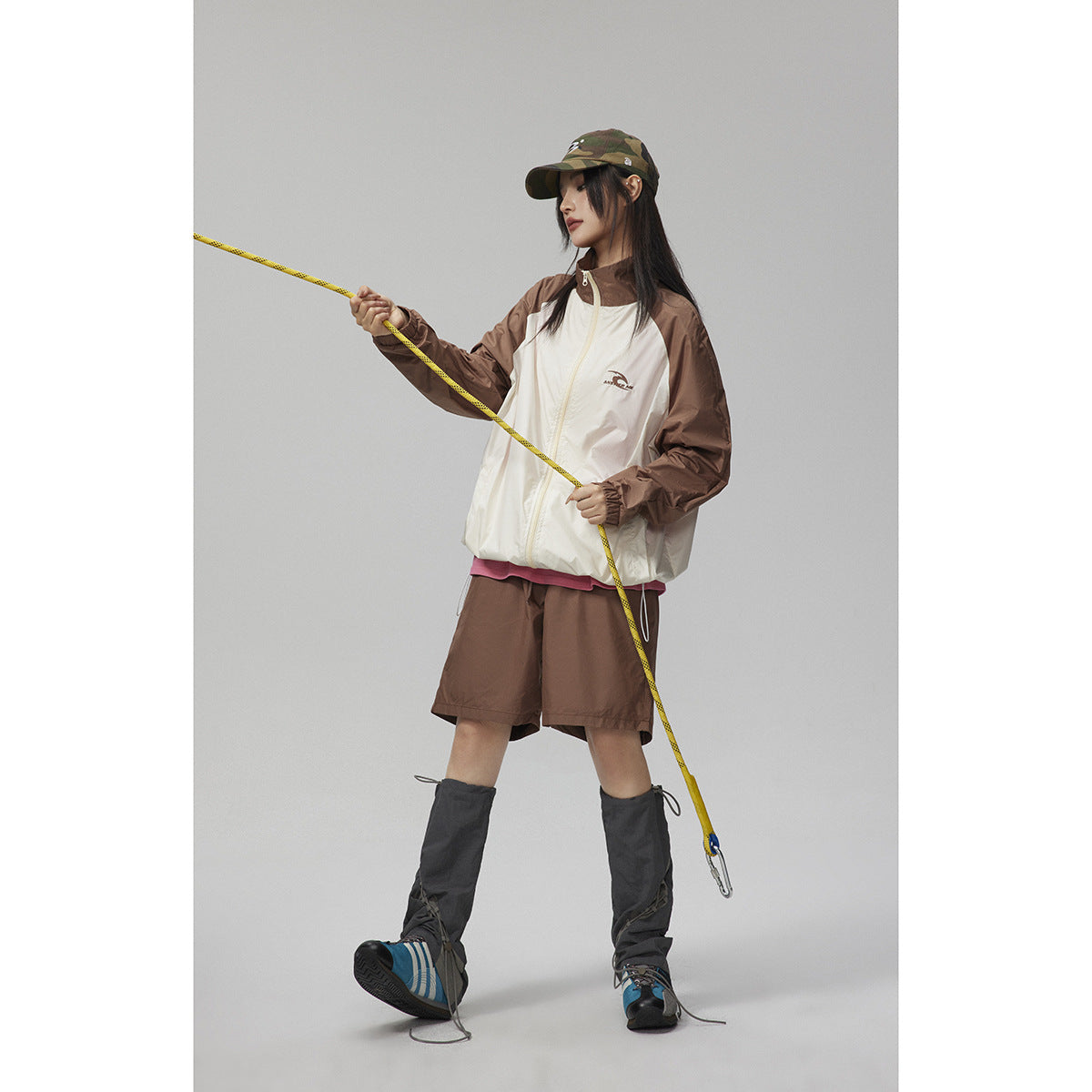 Model wearing a cream windbreaker with brown sleeves, brown shorts, a camouflage cap, and dark gaiters.