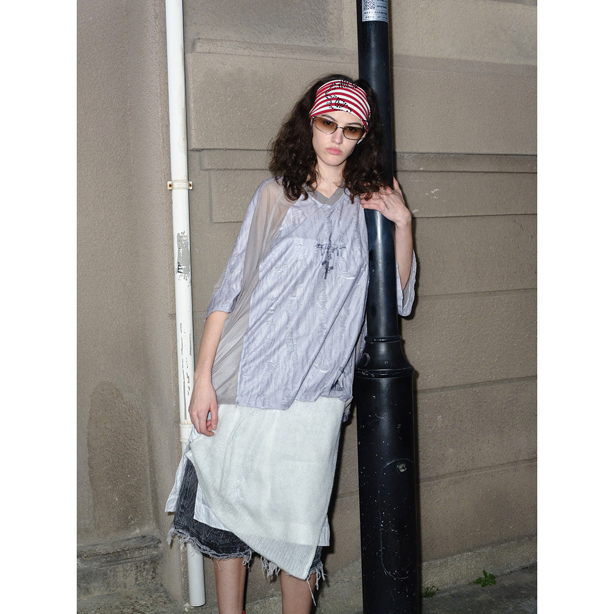 Model wearing a loose pale gray V-neck top with sheer taupe sleeves and a layered white skirt beside a street pole.