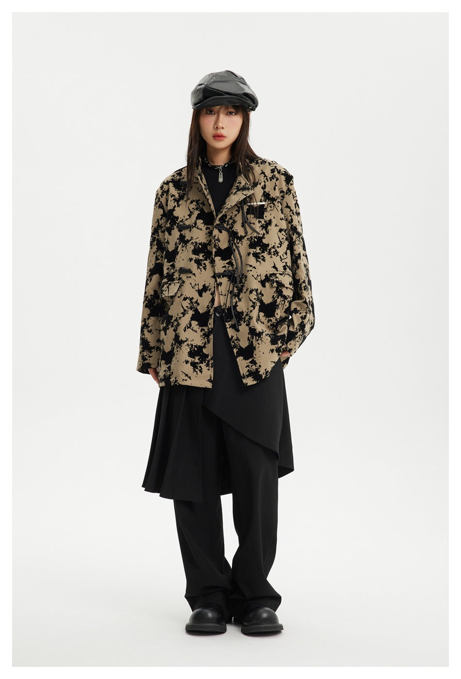 Model wearing khaki and black mottled stand-collar jacket with cord closures, black skirt layer, wide pants and a cap