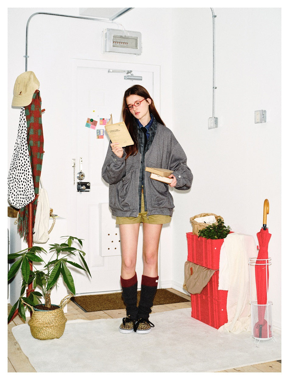 Full-length model wearing an oversized gray zip jacket over plaid layers and olive shorts, holding books in a room.
