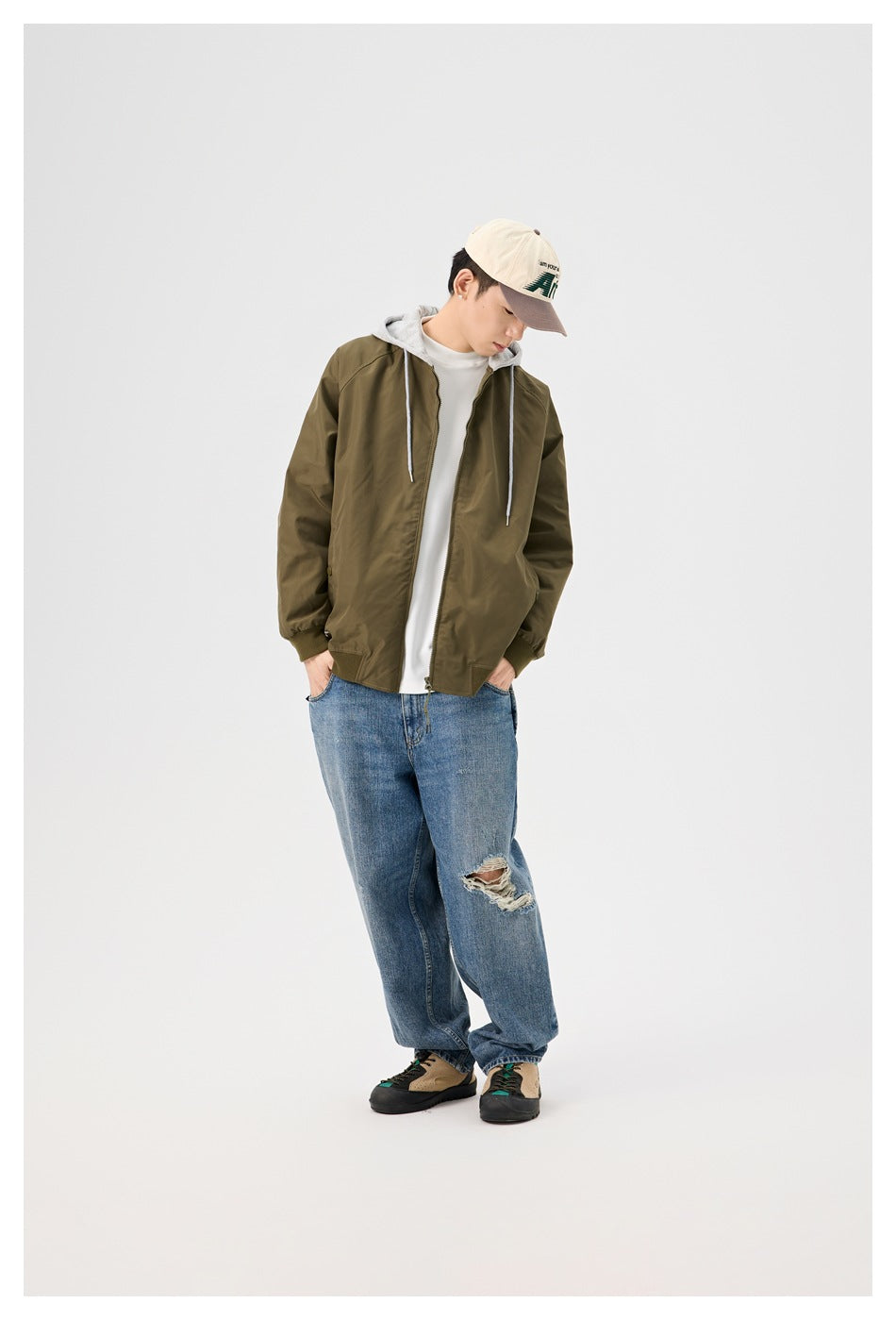 Model with head tilted down wearing the army green hooded zip jacket open over a white tee with blue jeans