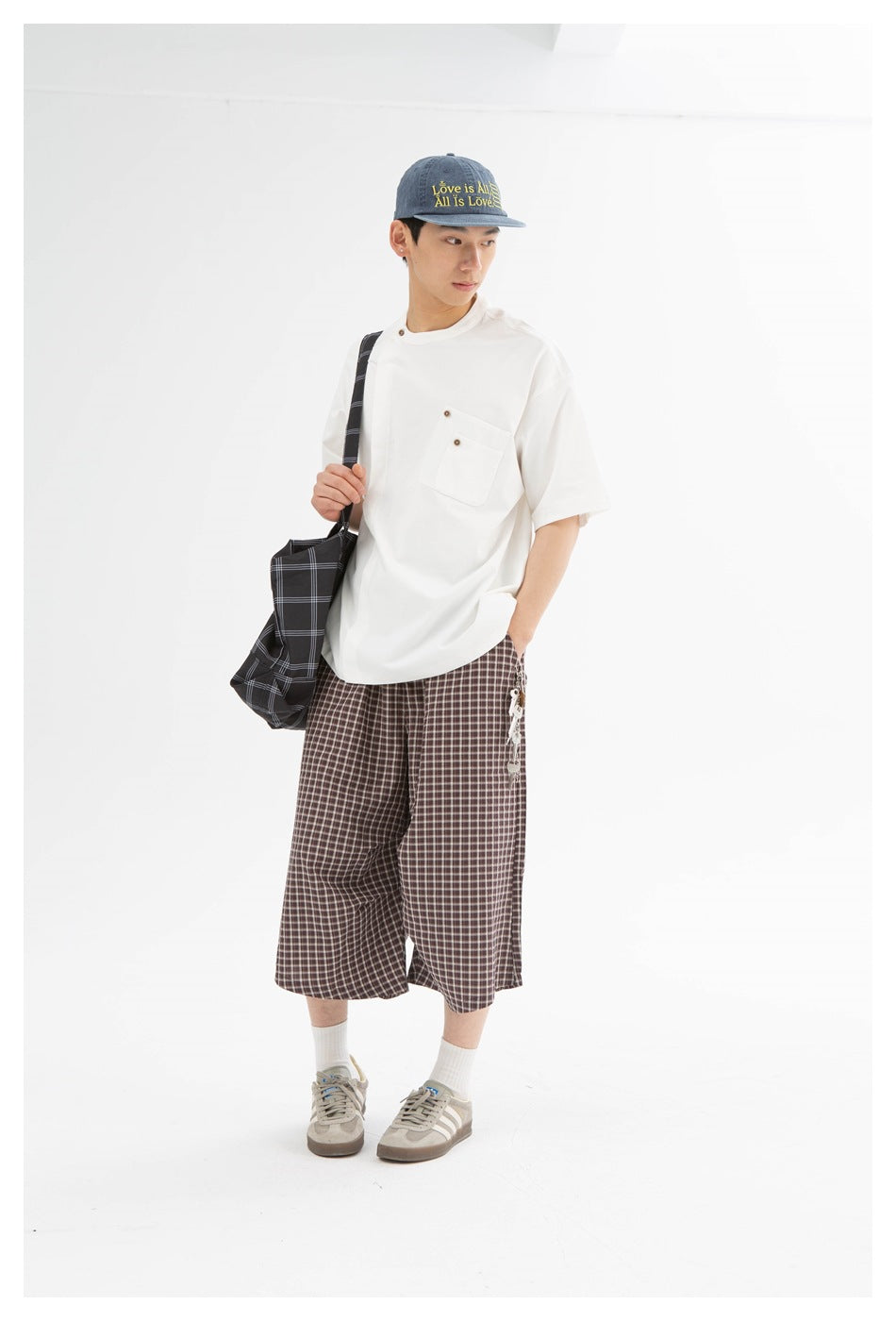 Man wearing a white short-sleeve pocket tee with checked cropped pants, a denim cap and a plaid tote bag