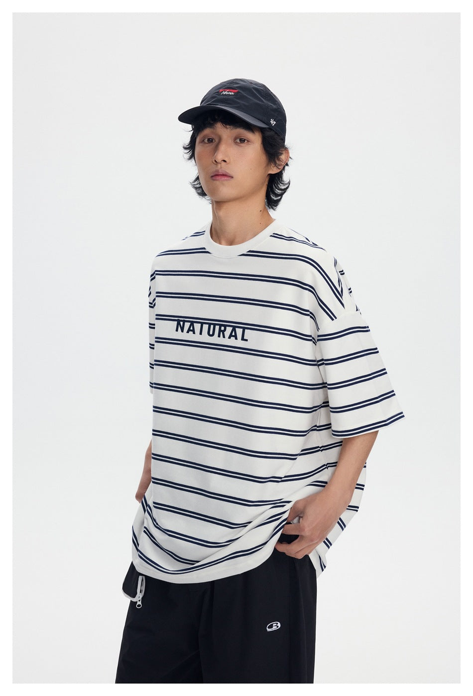 Model wearing white navy-striped oversized T-shirt with NATURAL print, angled view with cap and black pants