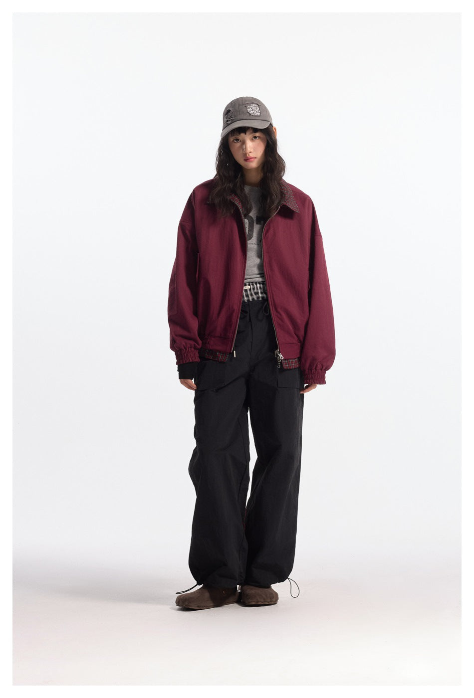 Model wearing open burgundy reversible jacket with plaid collar and hem, gray cap and black cargo pants