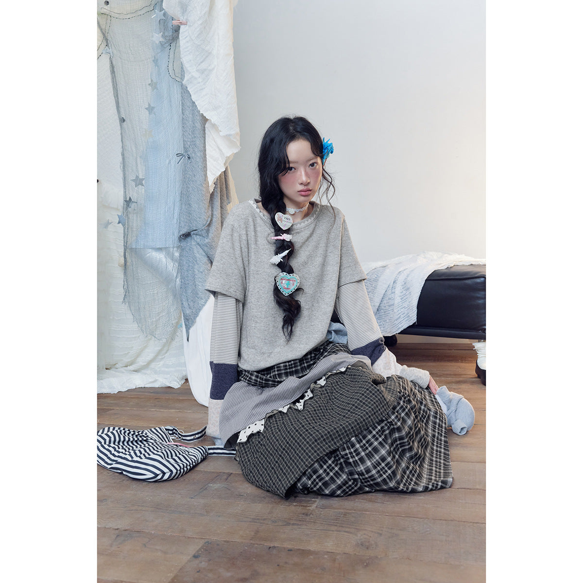Model seated in a gray short-sleeve top with striped patchwork sleeves and a long layered dark plaid skirt.