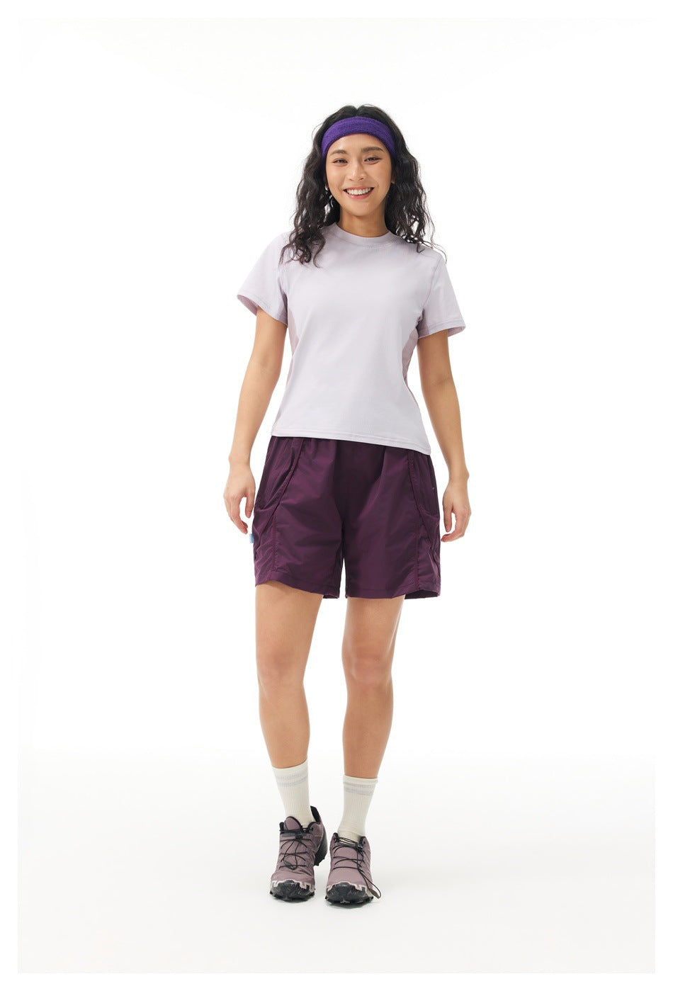Full-length front view of a model wearing a pale lavender T-shirt, plum shorts, and a purple headband.