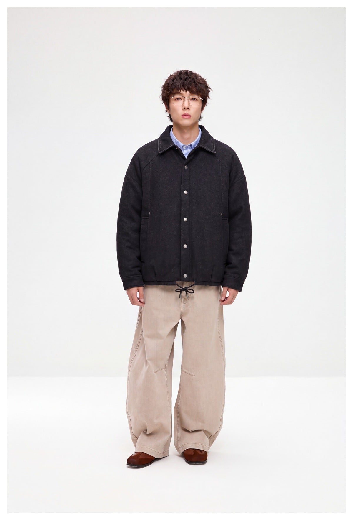 Full-length model wearing a black snap-front jacket with a pointed collar and beige trousers.