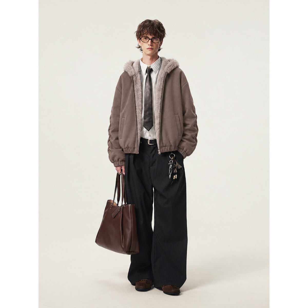 Man wearing a taupe hooded jacket with pale faux-fur trim, styled with a striped shirt and black trousers.