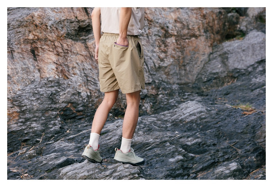 Rear three-quarter view of a model wearing tan shorts with back and side pockets outdoors.