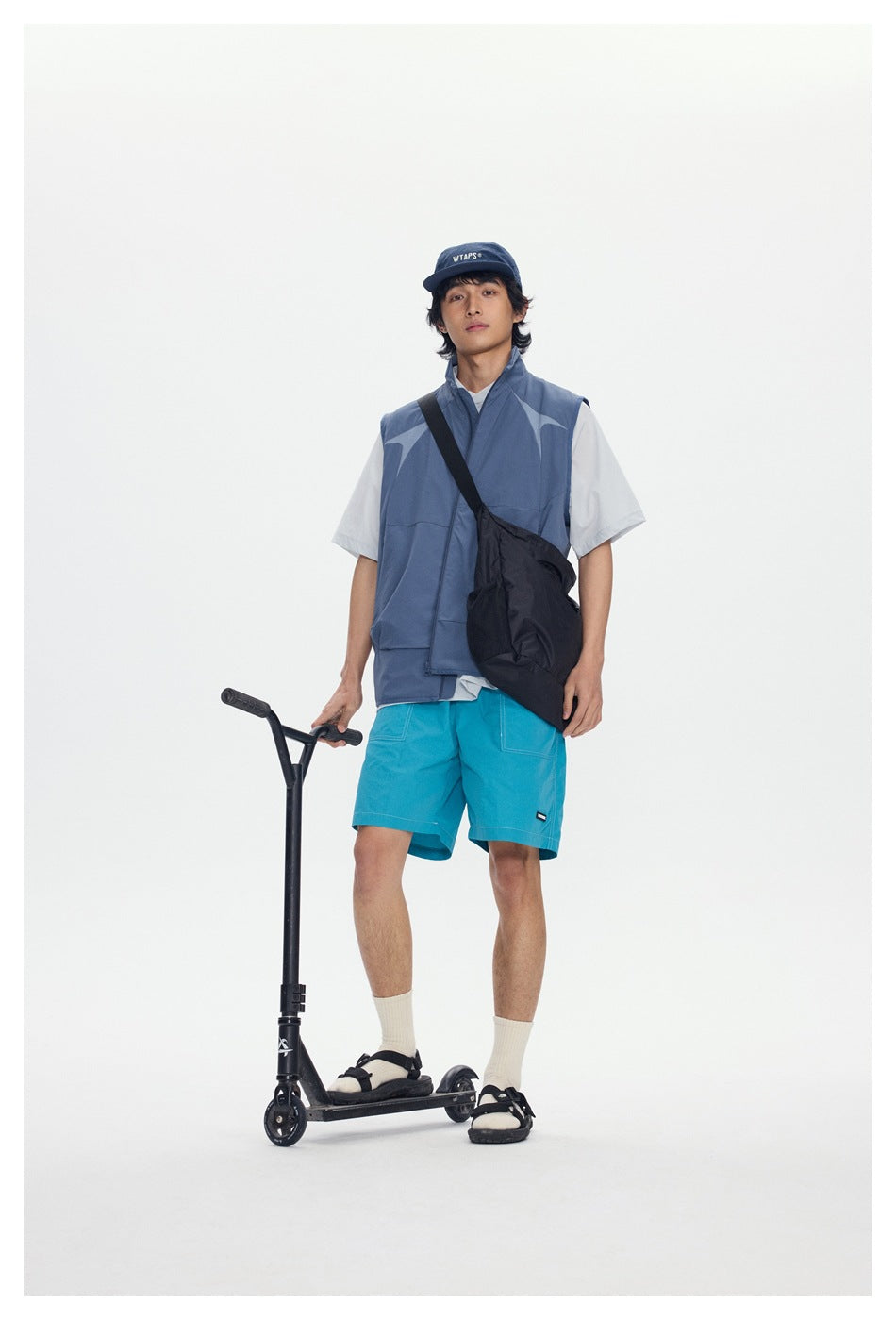Front view of a model wearing a blue-gray patterned vest, white T-shirt, turquoise shorts, and crossbody bag.
