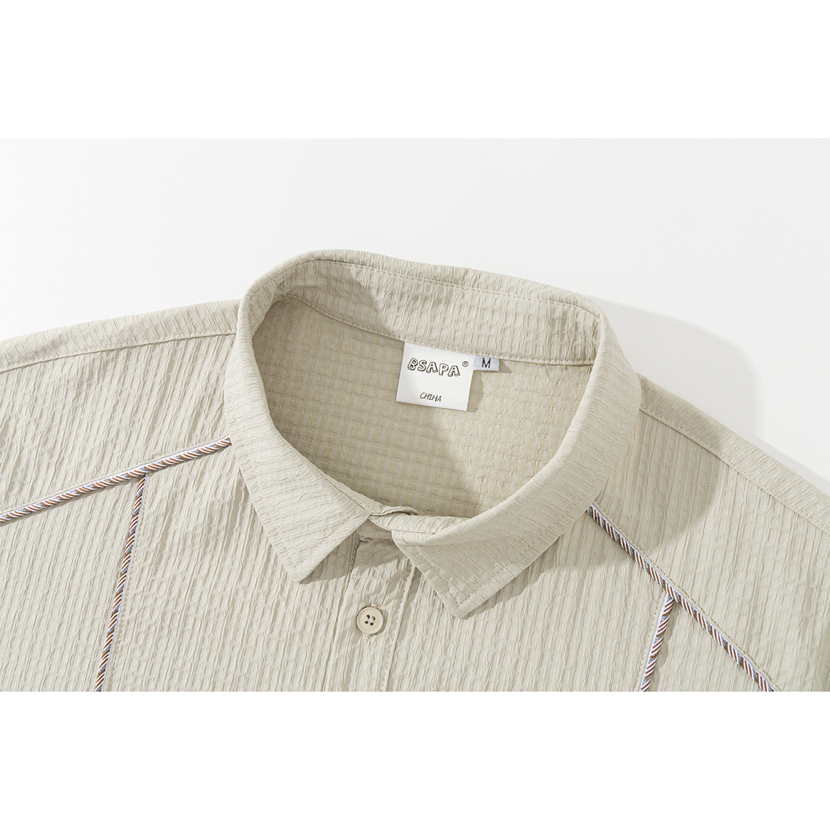 Close-up of a light beige shirt collar, textured fabric, button, contrast piping, and sewn-in neck label.