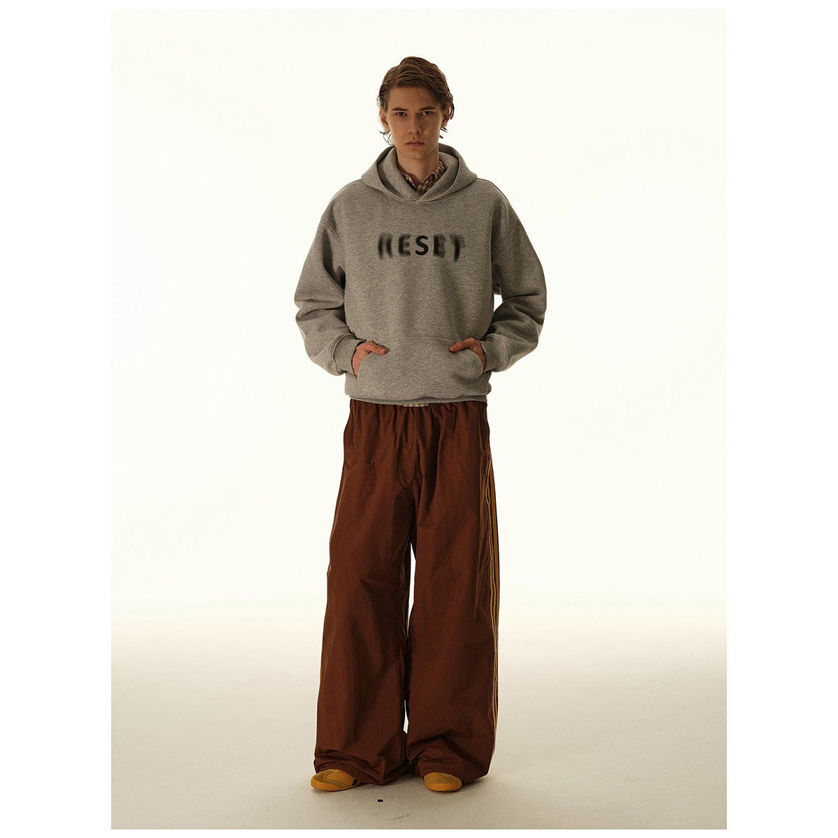 Model wearing a heather-gray hooded sweatshirt with dark chest lettering and wide brown trousers.