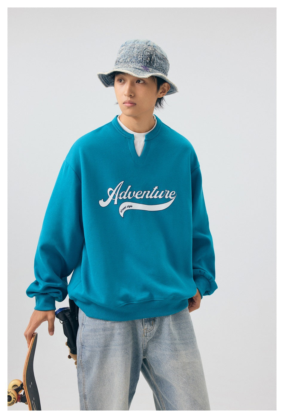 Model wearing a teal Adventure graphic sweatshirt with a notched neckline, shown from the front with jeans and a bucket hat.
