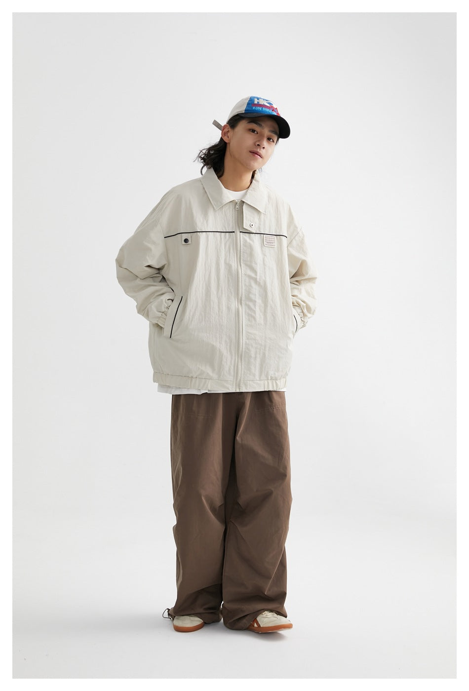 Full-length model wearing a zipped ivory jacket with black piping and loose brown trousers.