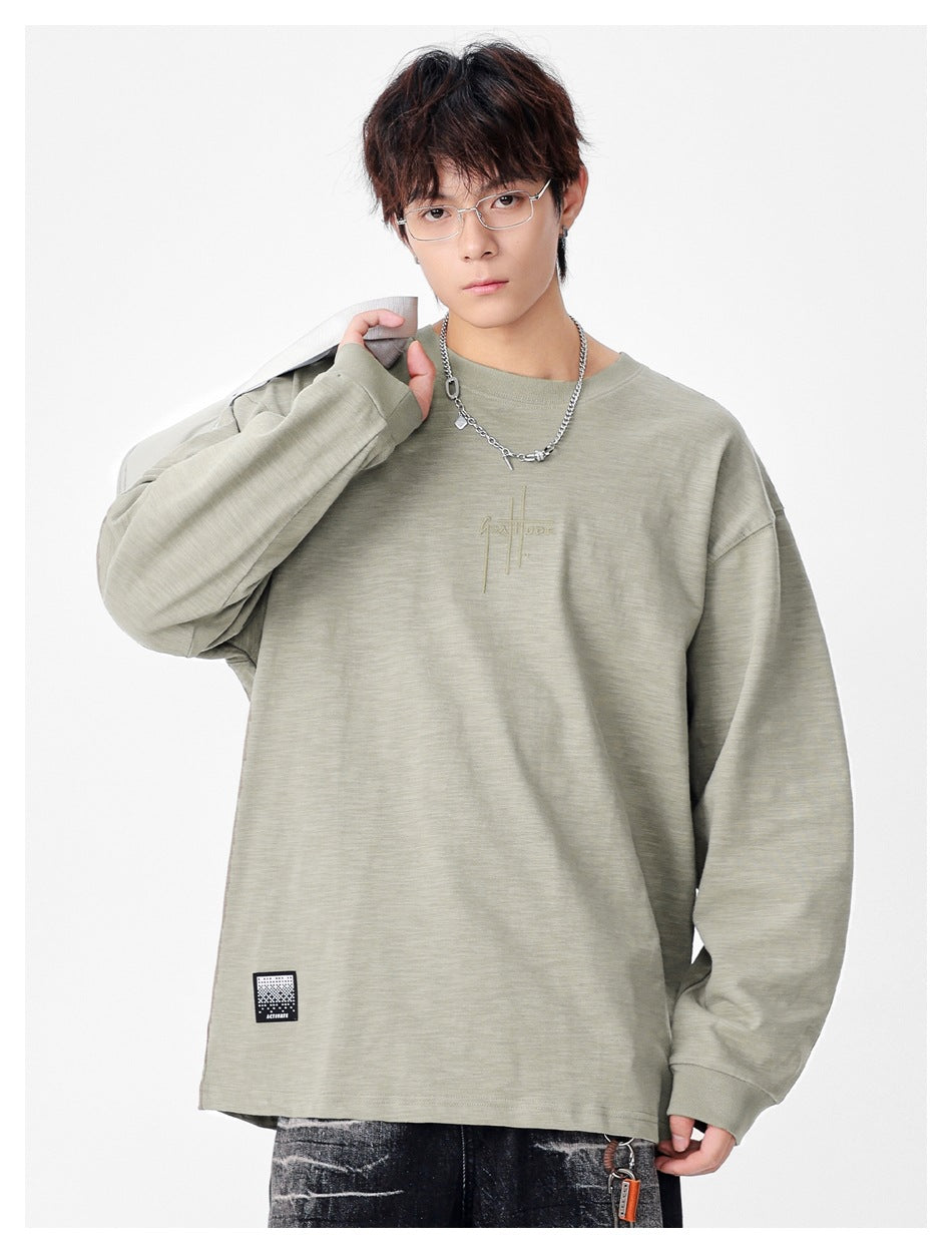 Model wearing a sage-gray oversized long-sleeve crewneck shirt with chest embroidery and a small hem patch.