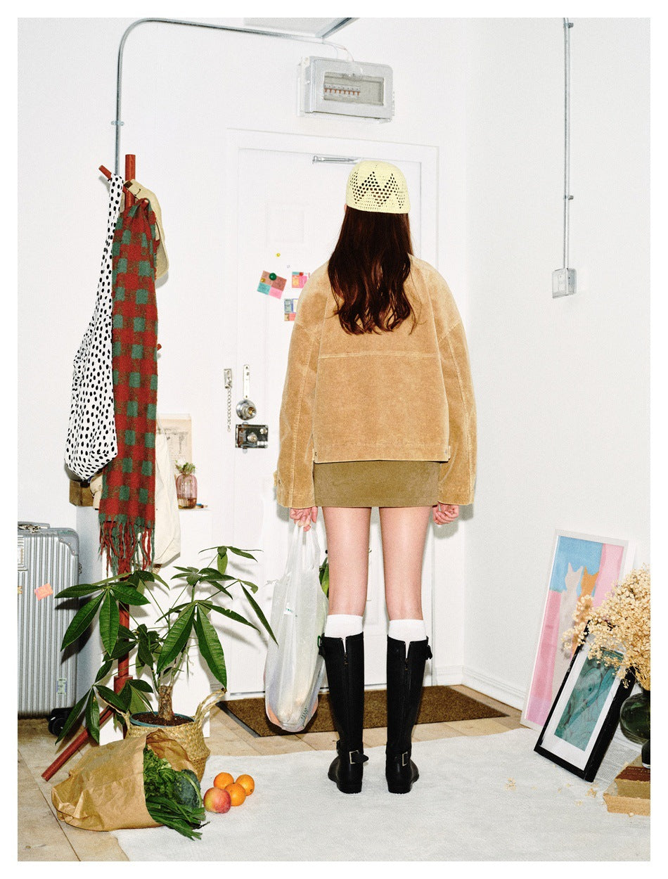 Rear view of a model wearing a tan cropped jacket and olive mini skirt with a cream knit cap and black knee-high boots.