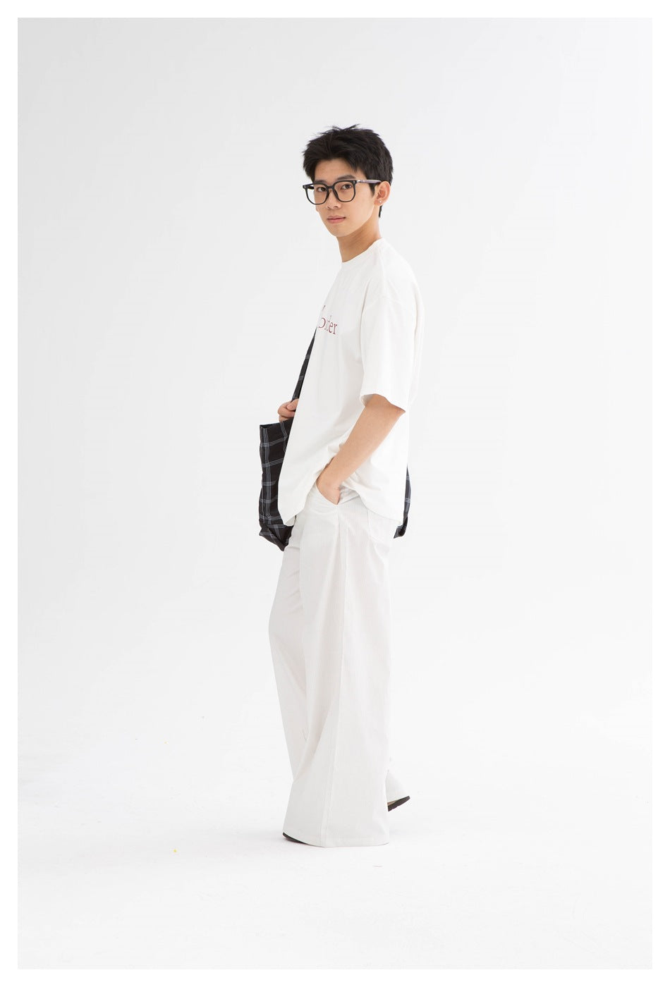 Side view of a model wearing a white oversized graphic T-shirt, wide-leg white pants, and a black plaid shoulder bag.