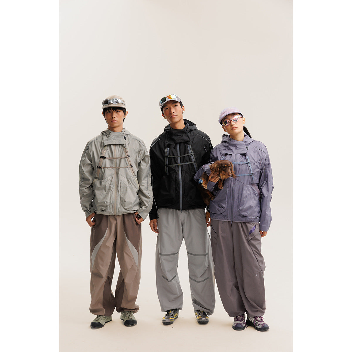 Three models wear gray, black, and lavender hooded jackets with loose outdoor pants; one holds a small dog.