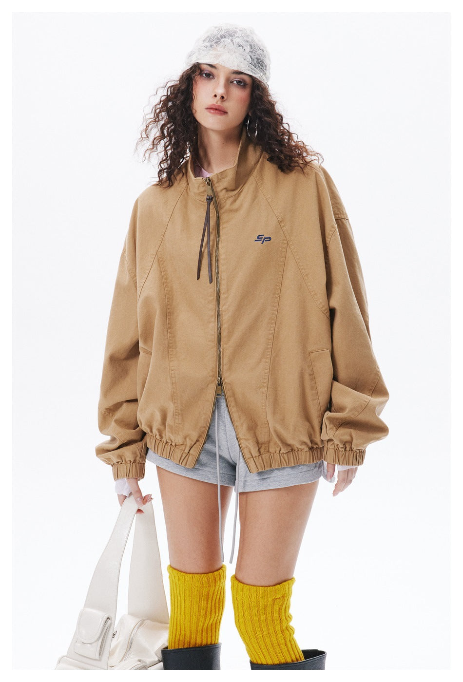 Closer front view of a model wearing an oversized tan jacket with a zip front, stand collar, and small chest emblem.