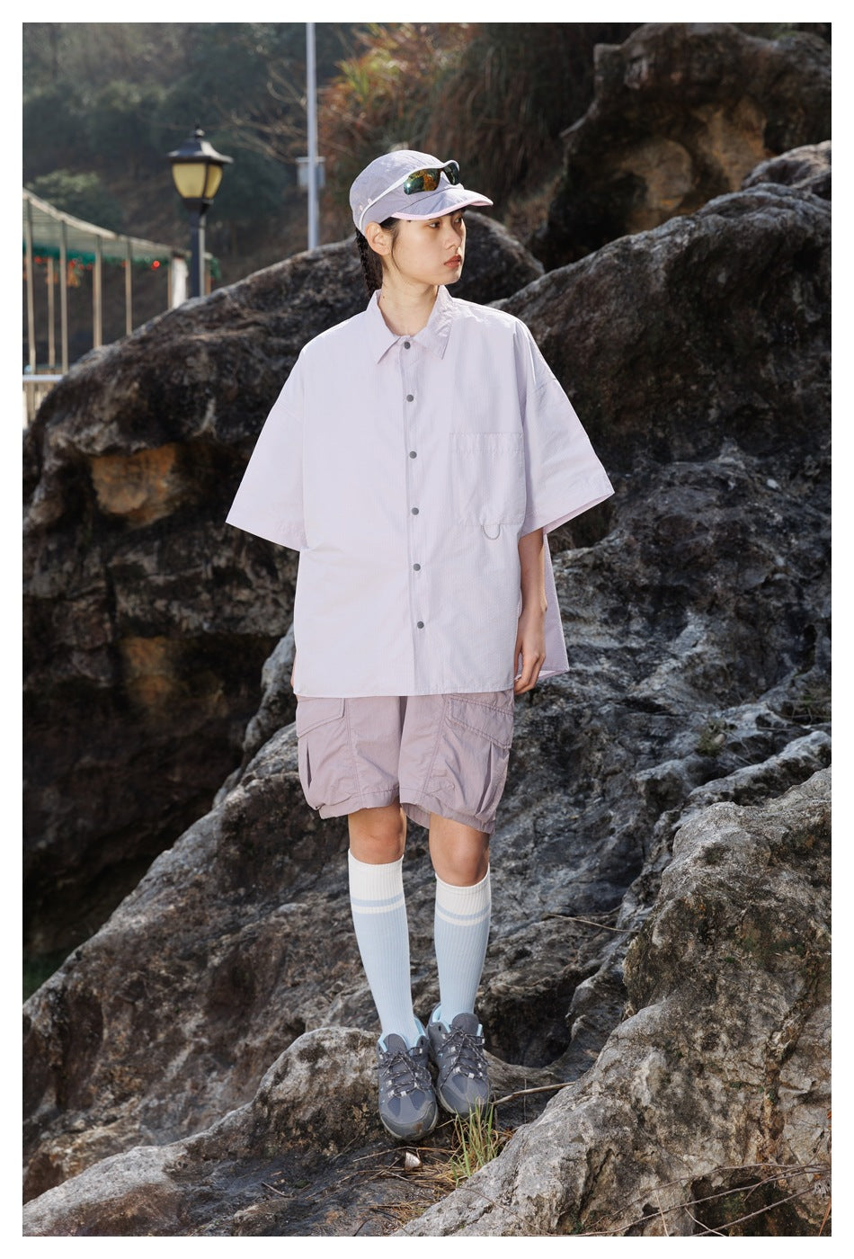 Model wearing an oversized pale lavender short-sleeve button-up shirt and mauve cargo shorts, shown outdoors.