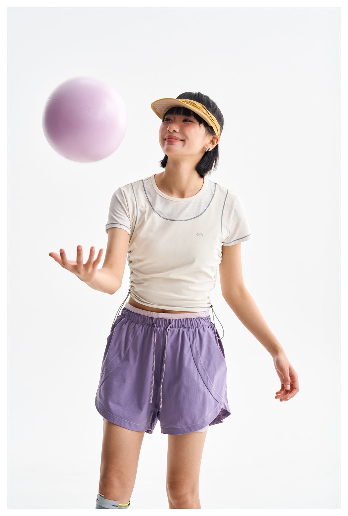 Model in a white contrast-seam athletic tee and lavender shorts, tossing a lavender ball.