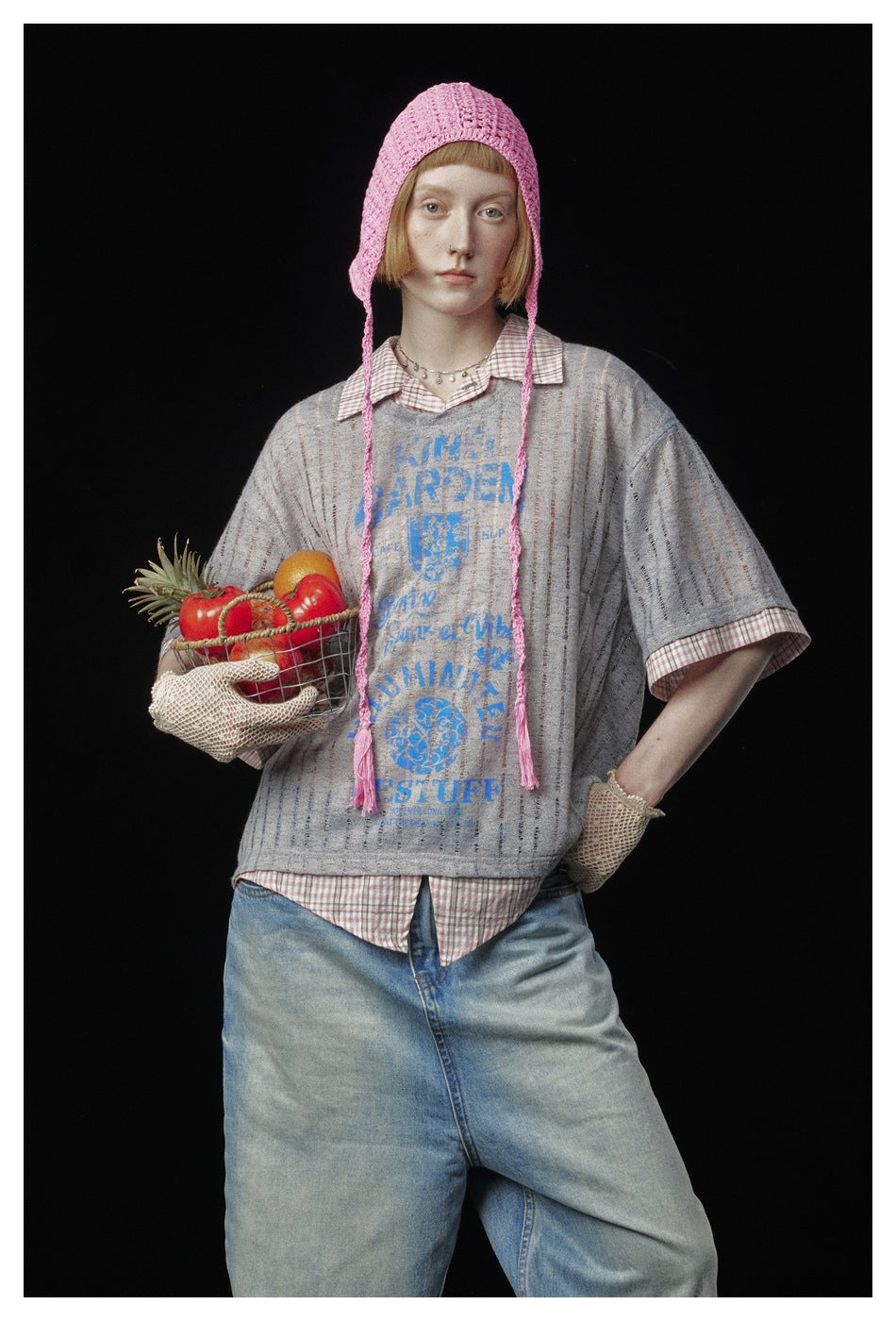 Model wearing a gray striped top with blue graphic over a checked shirt, paired with a pink crocheted bonnet and gloves.