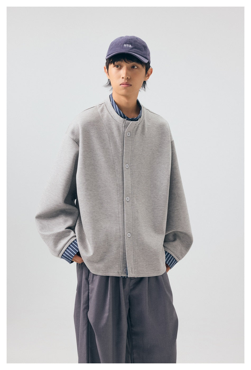 Model wearing a light gray button-front jacket over a striped shirt, with dark trousers and a purple cap.