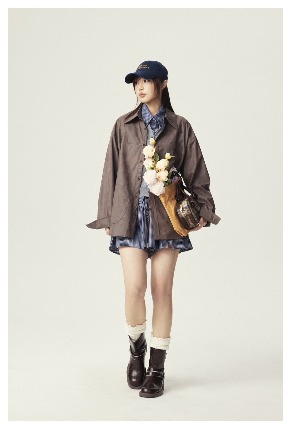 Model wearing an oversized brown button-up jacket and blue checked outfit, accessorized with a cap and carrying flowers and