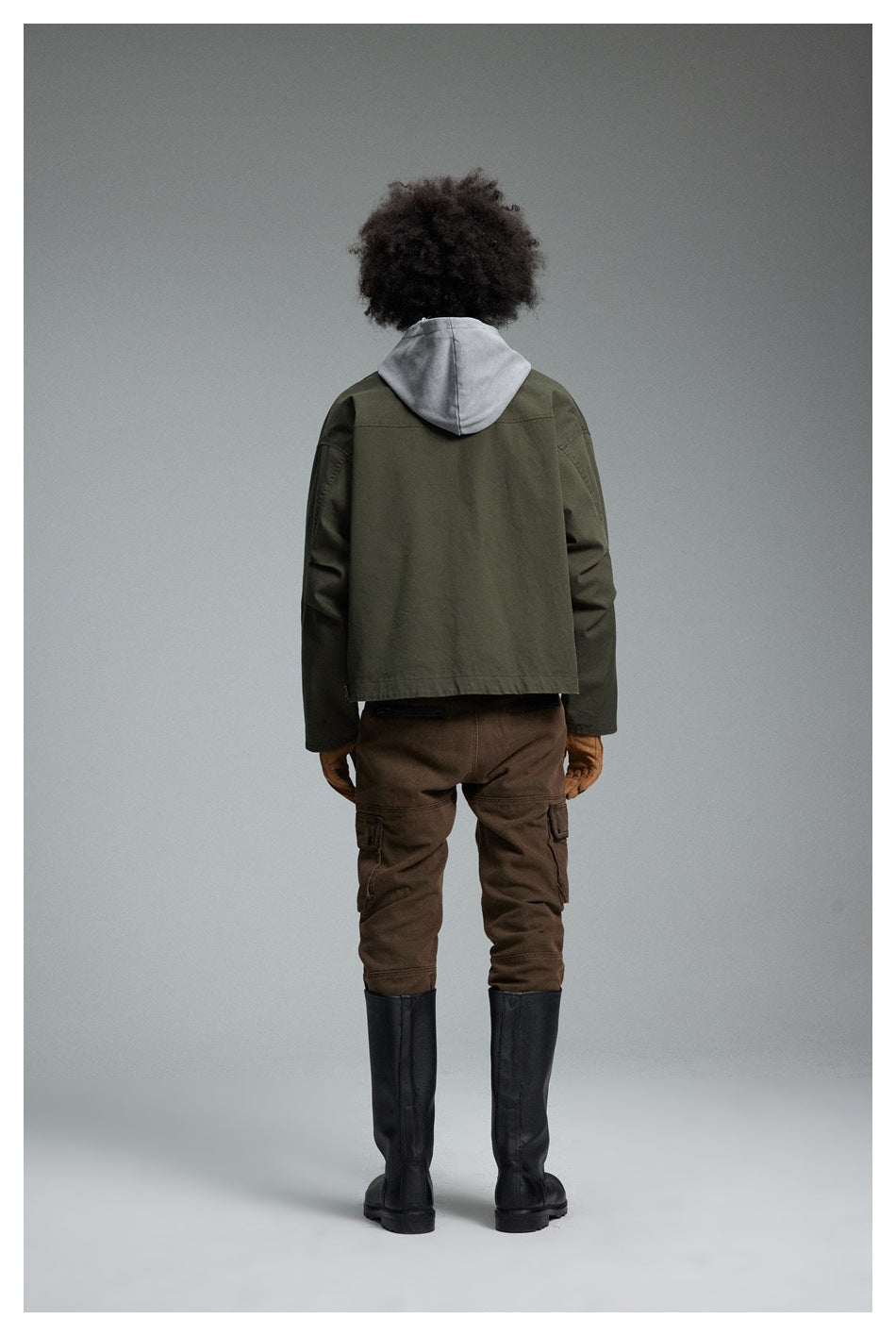 Back view of a model wearing the army green jacket over a gray hood with brown pants and black tall boots