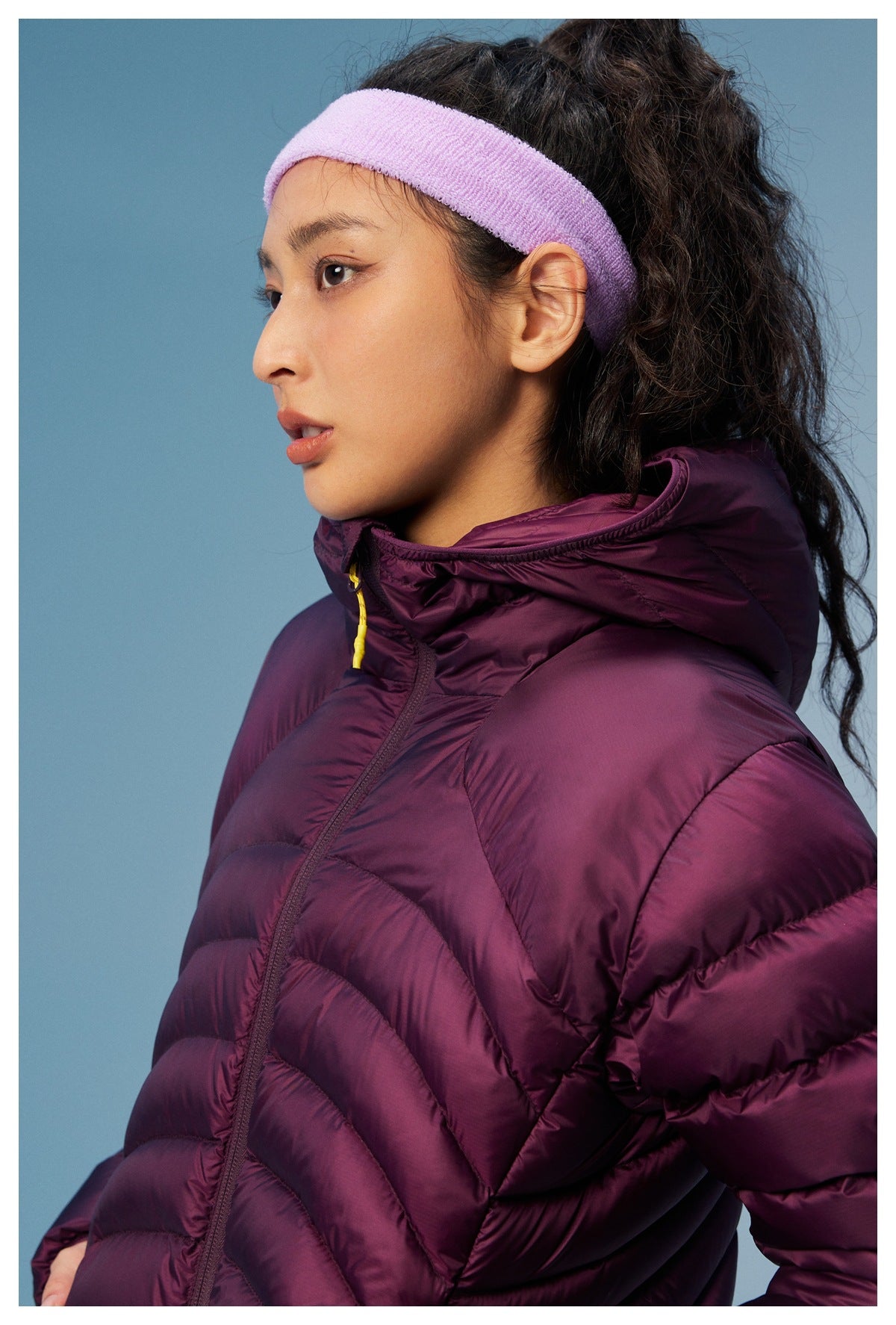 Close-up of the plum quilted jacket's hood, high collar, zipper, and yellow pull beside a lavender headband.