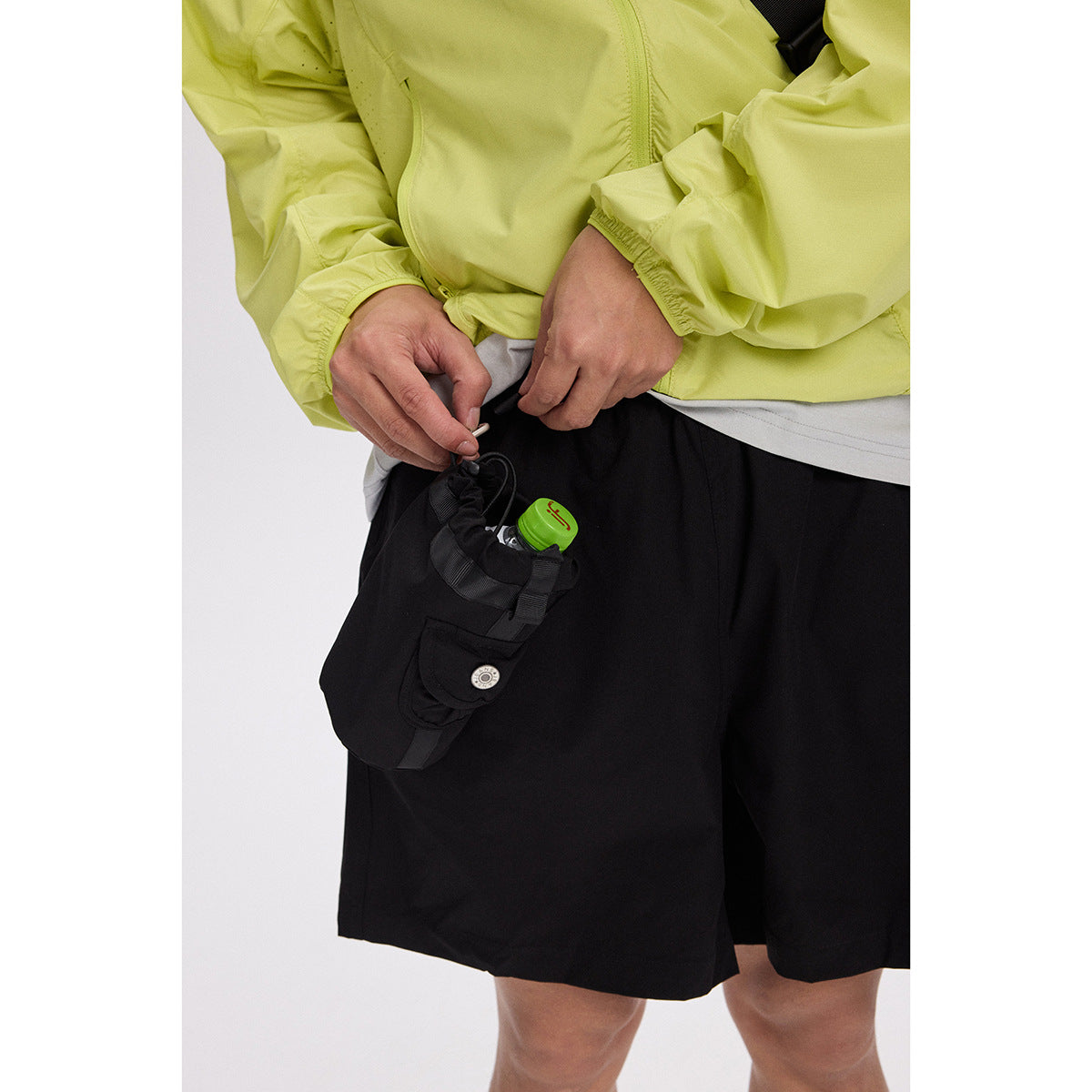 Close-up of a person wearing black shorts and a lime jacket, adjusting a small utility pouch attached to the shorts.