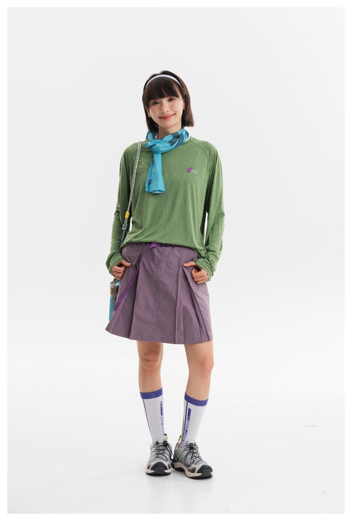 Full-length model wearing a green long-sleeve shirt, lavender pleated skirt, blue scarf and white knee socks.