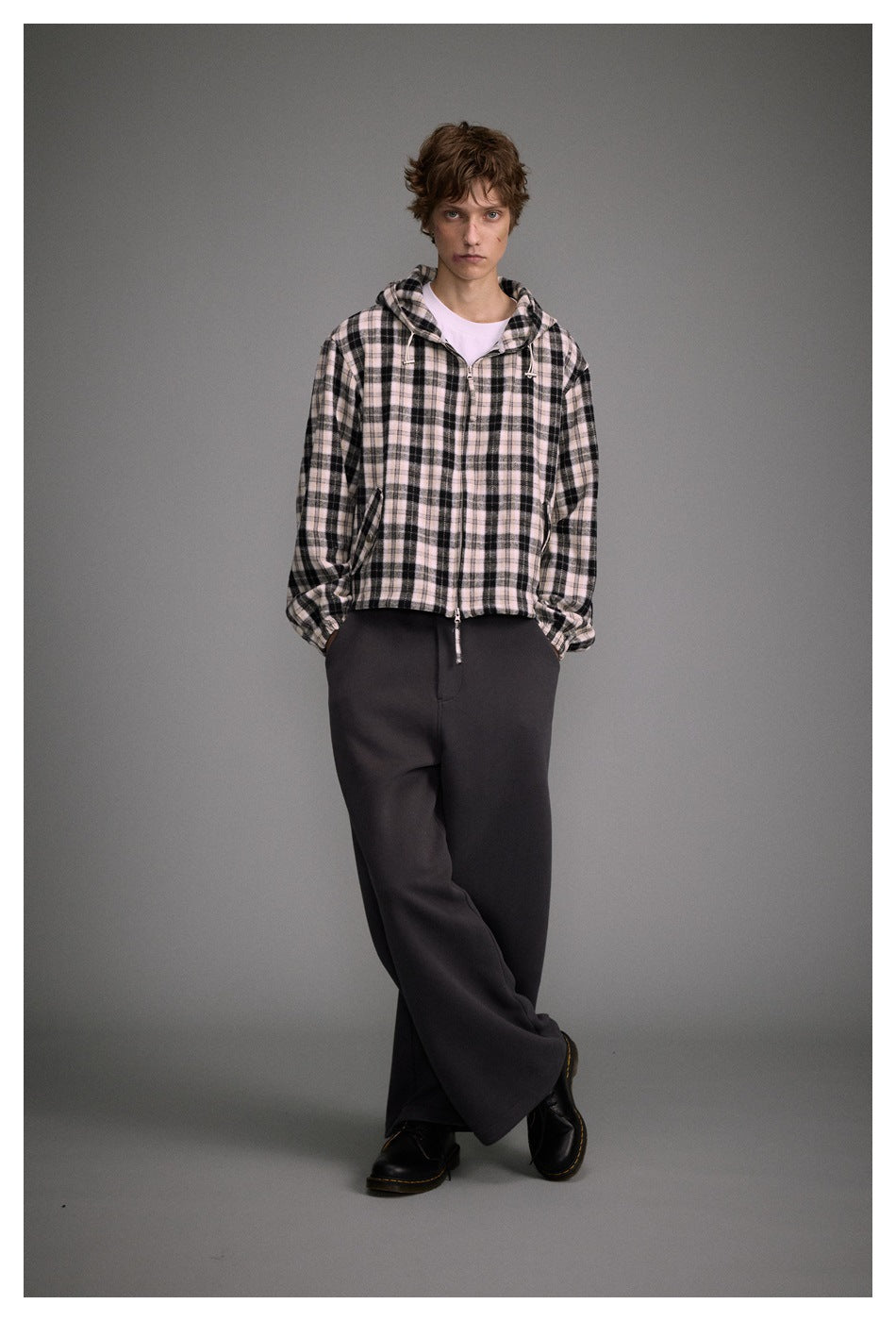 Full-length model wearing a cream and black plaid hooded zip jacket with dark trousers.
