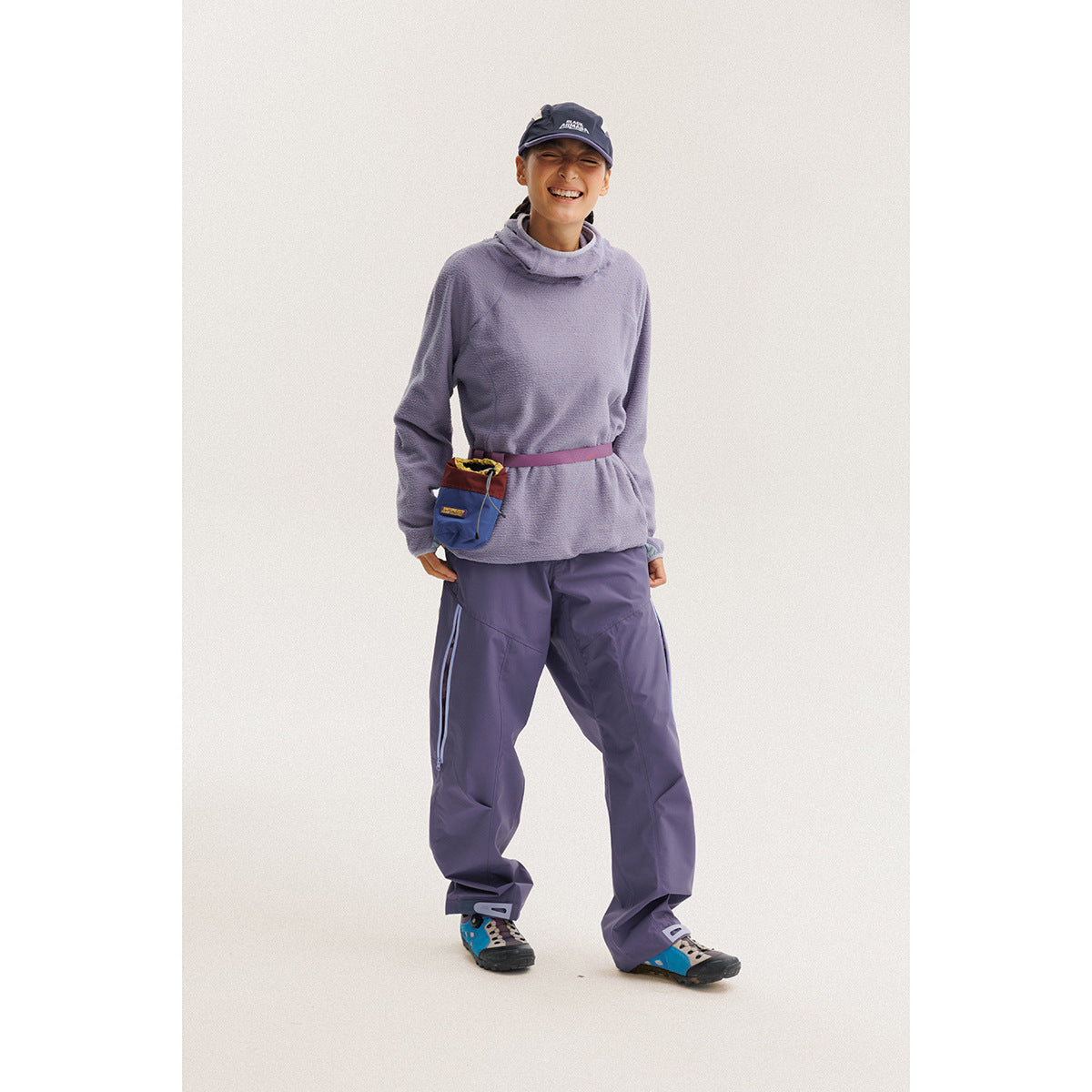 Model wearing a lavender cowl-neck fleece pullover, purple pants, a navy cap, and a small waist pouch.
