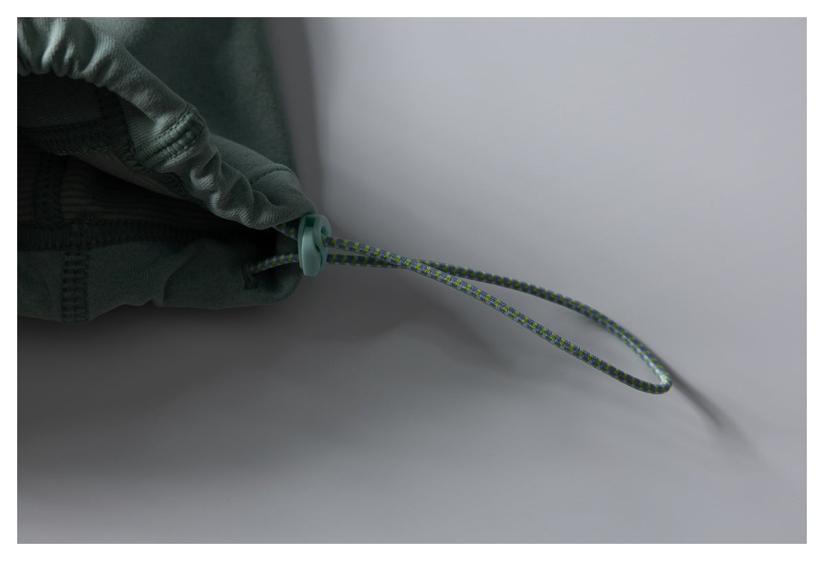 Close-up of a jacket hem drawcord and toggle against sage-green fabric.