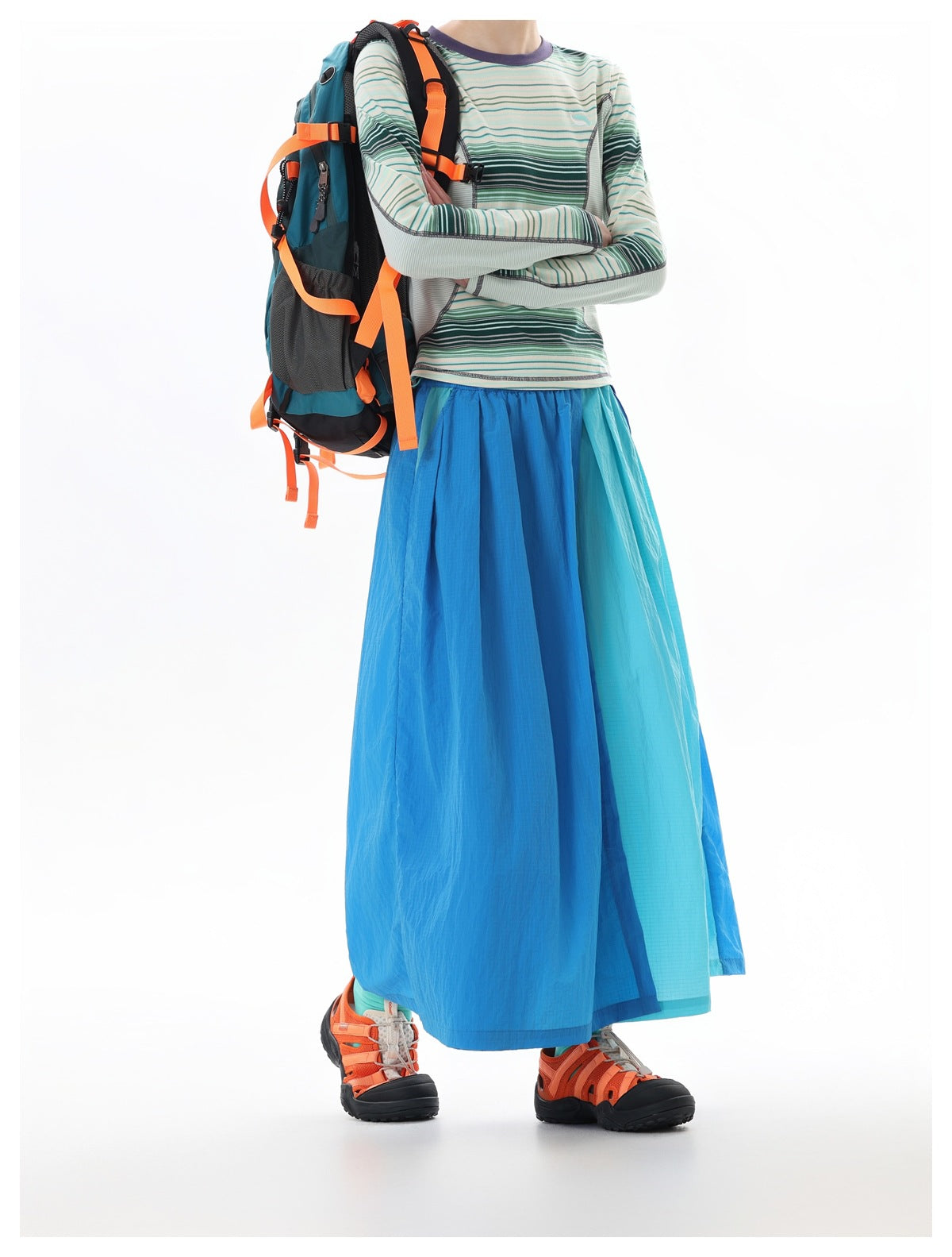 Model in a blue and aqua color-block maxi skirt and striped long-sleeve top, carrying a teal backpack with orange straps.