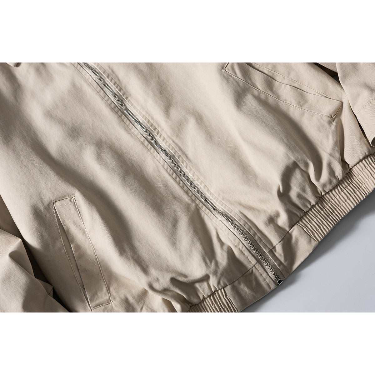 Close-up of a beige jacket front showing the silver zipper, welt pockets and gathered elastic hem.