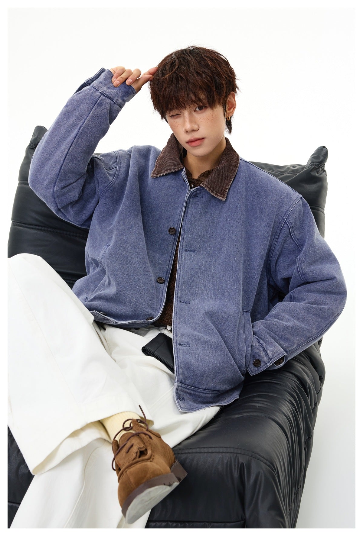 Seated model in a faded blue button-front jacket with a brown collar, styled with white trousers.
