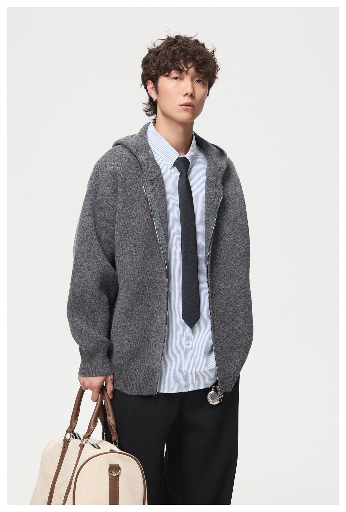 Close view of a man in a dark gray knit hooded zip jacket over a striped shirt and tie, carrying a tote bag