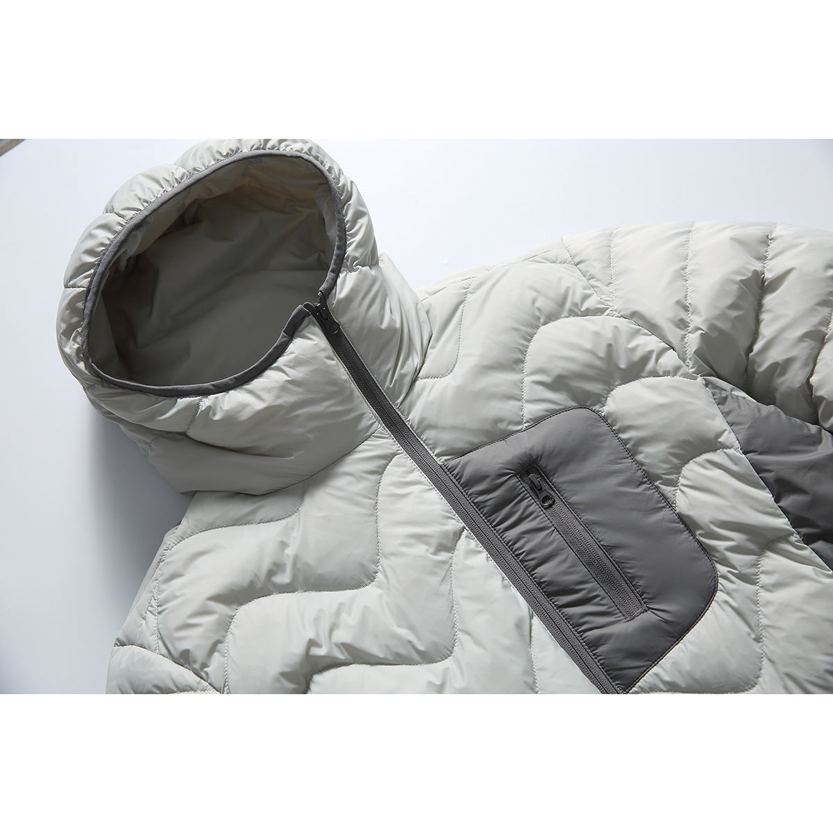 Close-up of a light gray hooded puffer jacket with dark gray trim, a diagonal front zipper, and a zippered chest pocket.