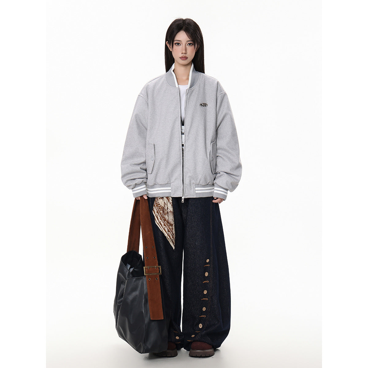 Full-length model wearing a light gray bomber jacket with striped ribbing, wide dark jeans, and carrying a large black tote.