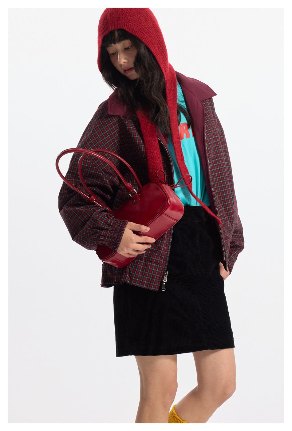 Model wearing an open red-and-green plaid jacket with burgundy panels and a red knit hood, shown with a red handbag.